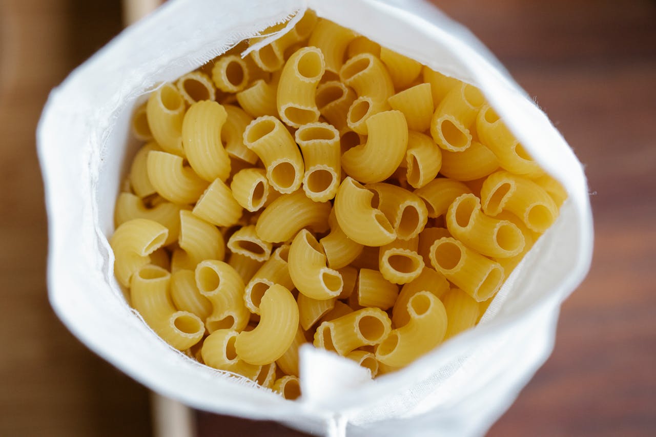 services-04 Top view white eco friendly sack full of uncooked macaroni placed on table on blurred background in light kitchen at home