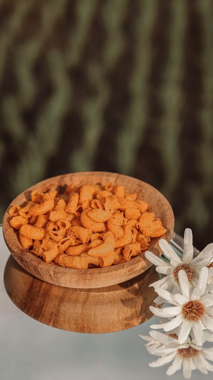 why-choose-us Close-up of a wooden bowl filled with savory Indonesian snacks on a reflective surface beside daisy flowers.